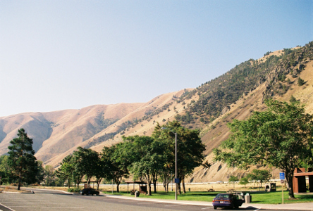 [The rest stop and highway are in the valley of the canyon. There are trees growing at the rest stop, but they are dwarfed by the canyon hills. The hills are mostly brown, but do have some tree'd sections.]